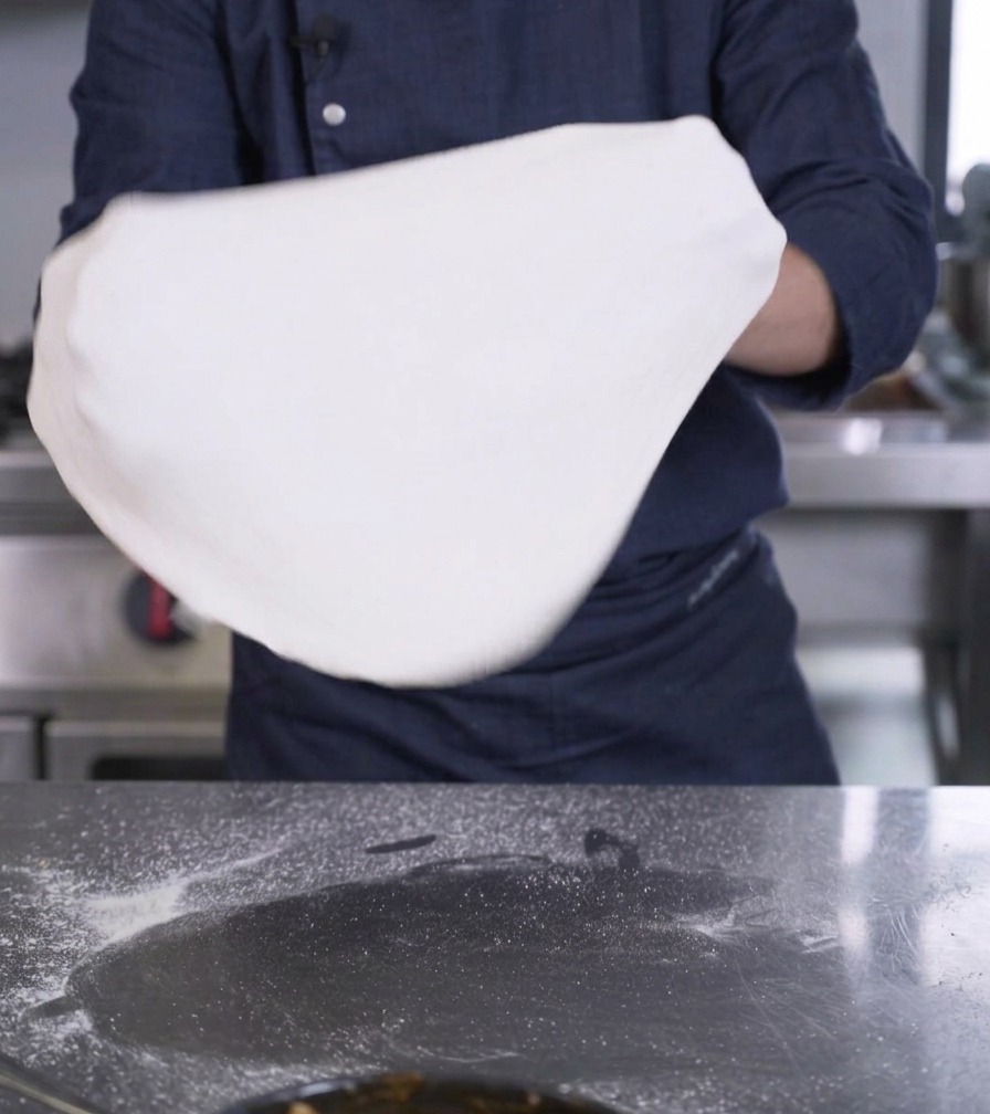 A persons hands holding up and stretching a large, thin disk of white pizza dough.