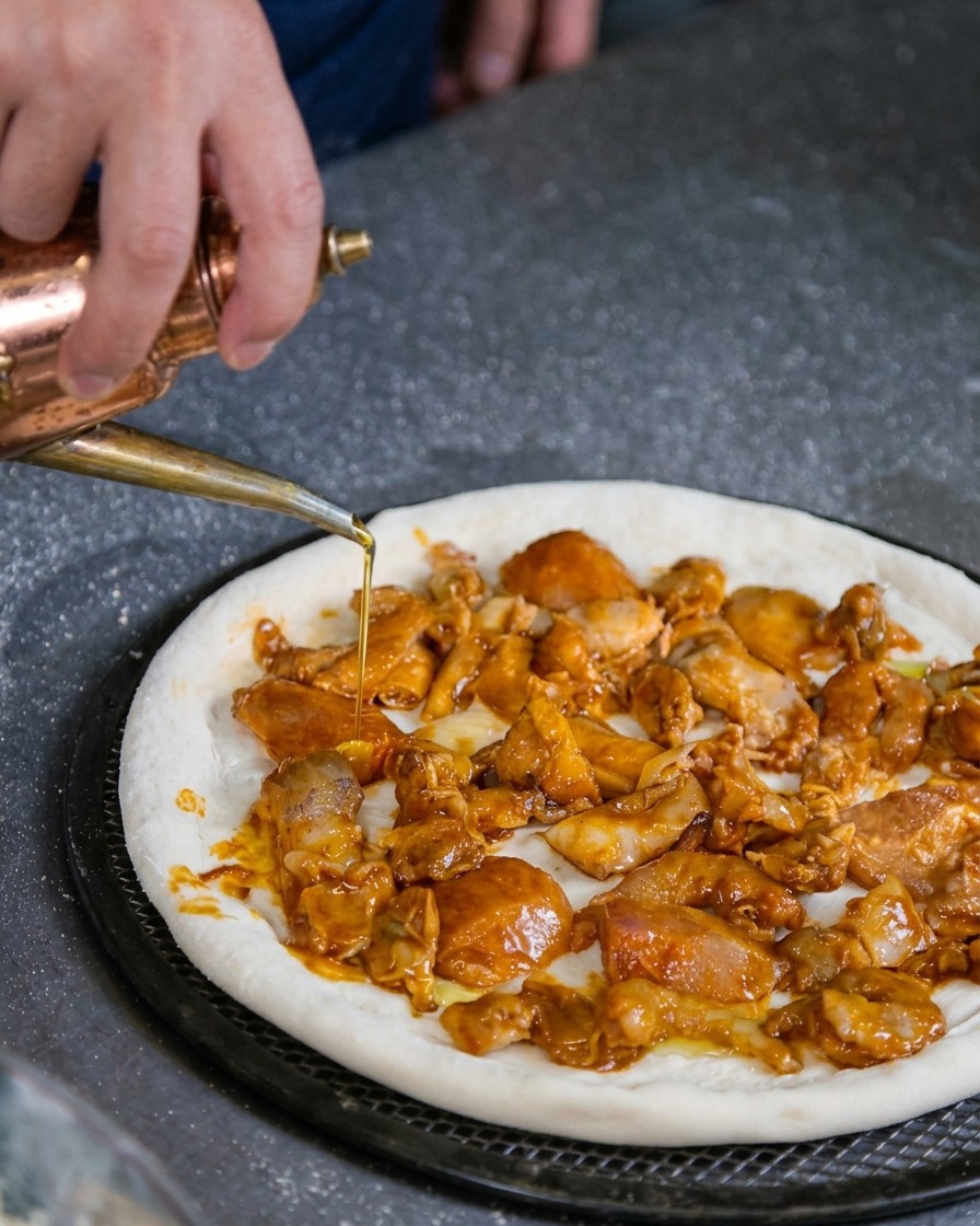 A hand using a copper oil dispenser to gently drizzle olive oil over an unbaked pizza base covered with a marinated seafood mixture.
