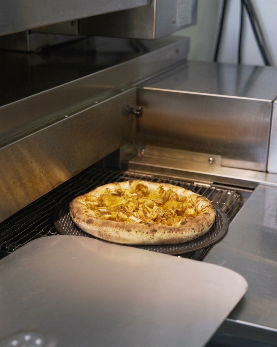 A seafood pizza moving through a commercial conveyor oven with its crust puffing up and browning.