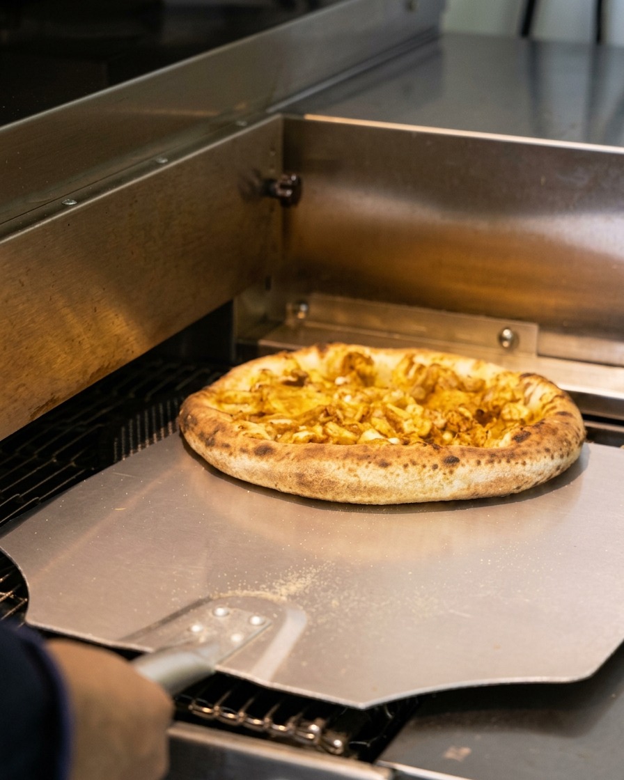 A freshly baked seafood pizza with a puffed, slightly charred crust being pulled from a commercial oven using a metal pizza peel.