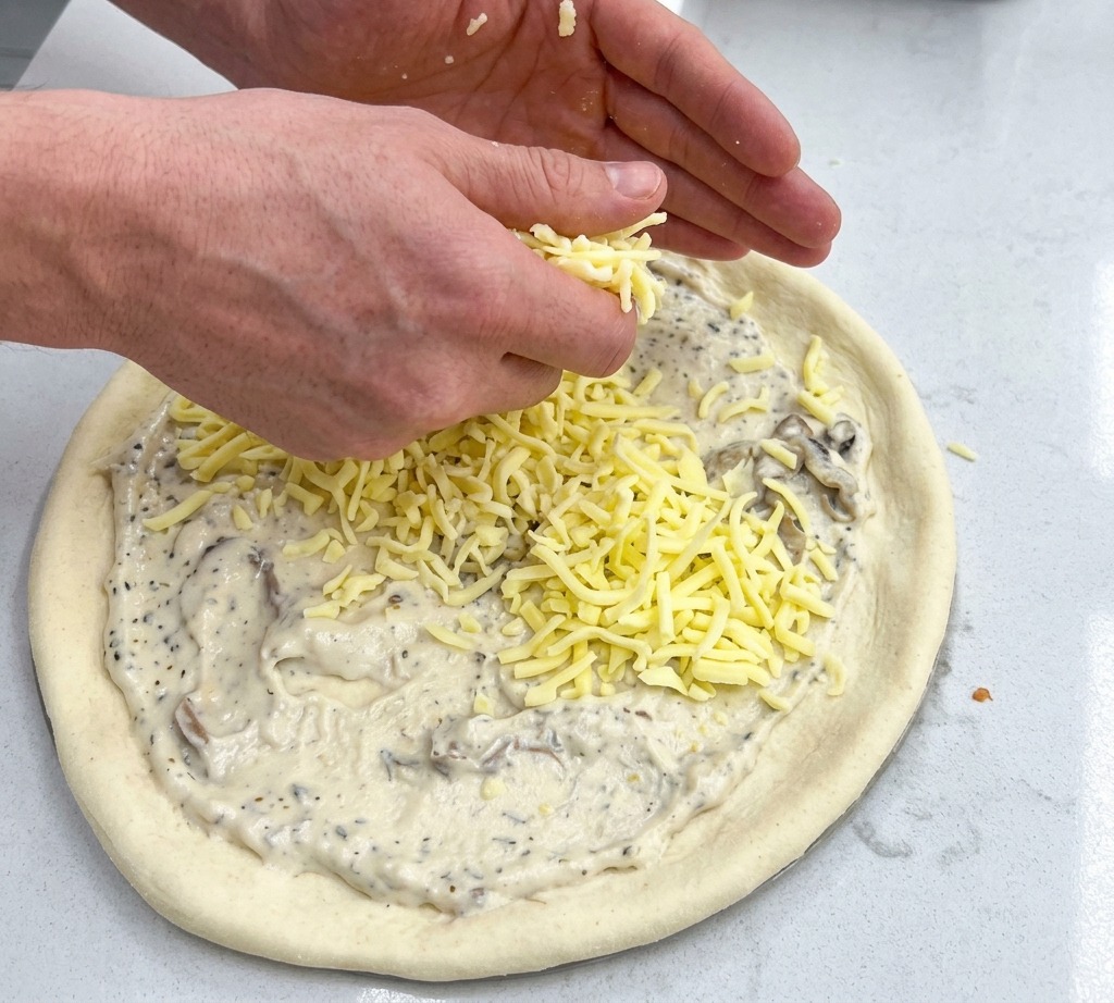 Two hands sprinkling handfuls of shredded yellow mozzarella cheese over a pizza base coated in cream sauce.