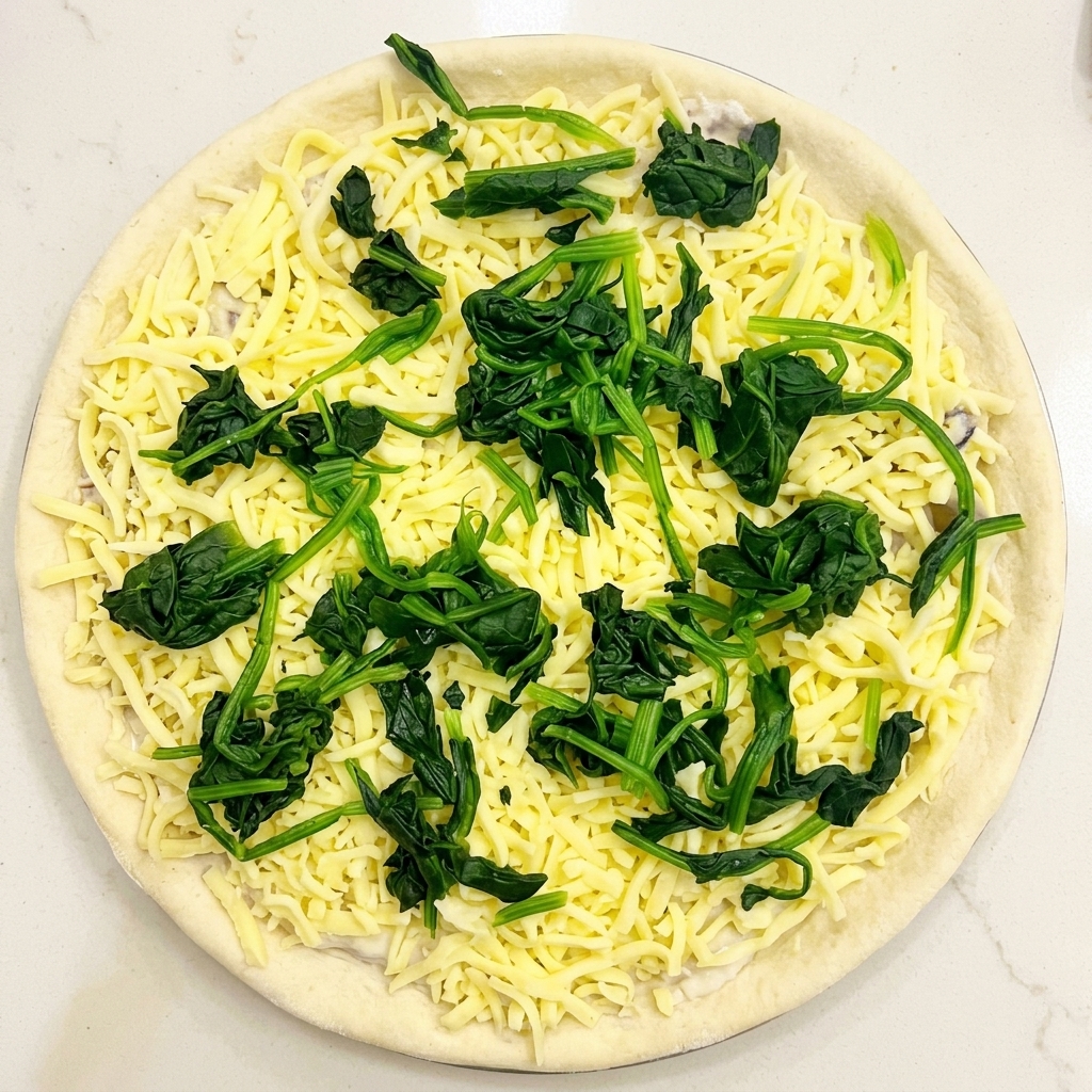 Top-down view of raw pizza dough topped with cream sauce, shredded mozzarella, and scattered portions of blanched green spinach.