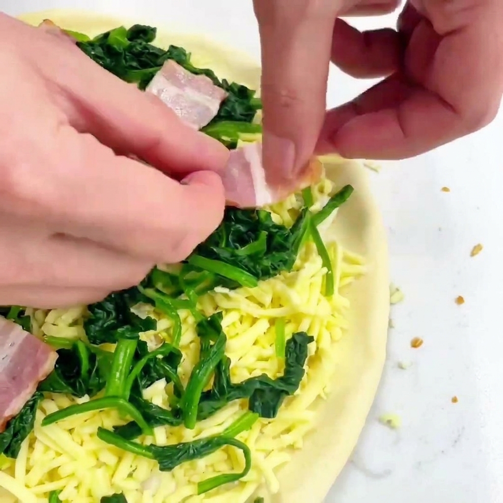 Hands placing square cuts of pink raw bacon onto a pizza topped with shredded cheese and green spinach.