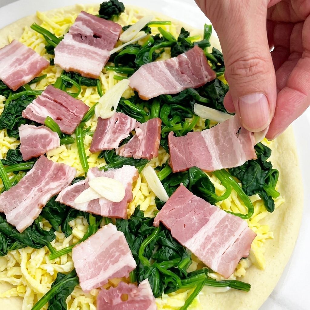 Hand dropping thin slices of raw garlic over an uncooked pizza completely covered with cheese, spinach, and raw bacon squares.