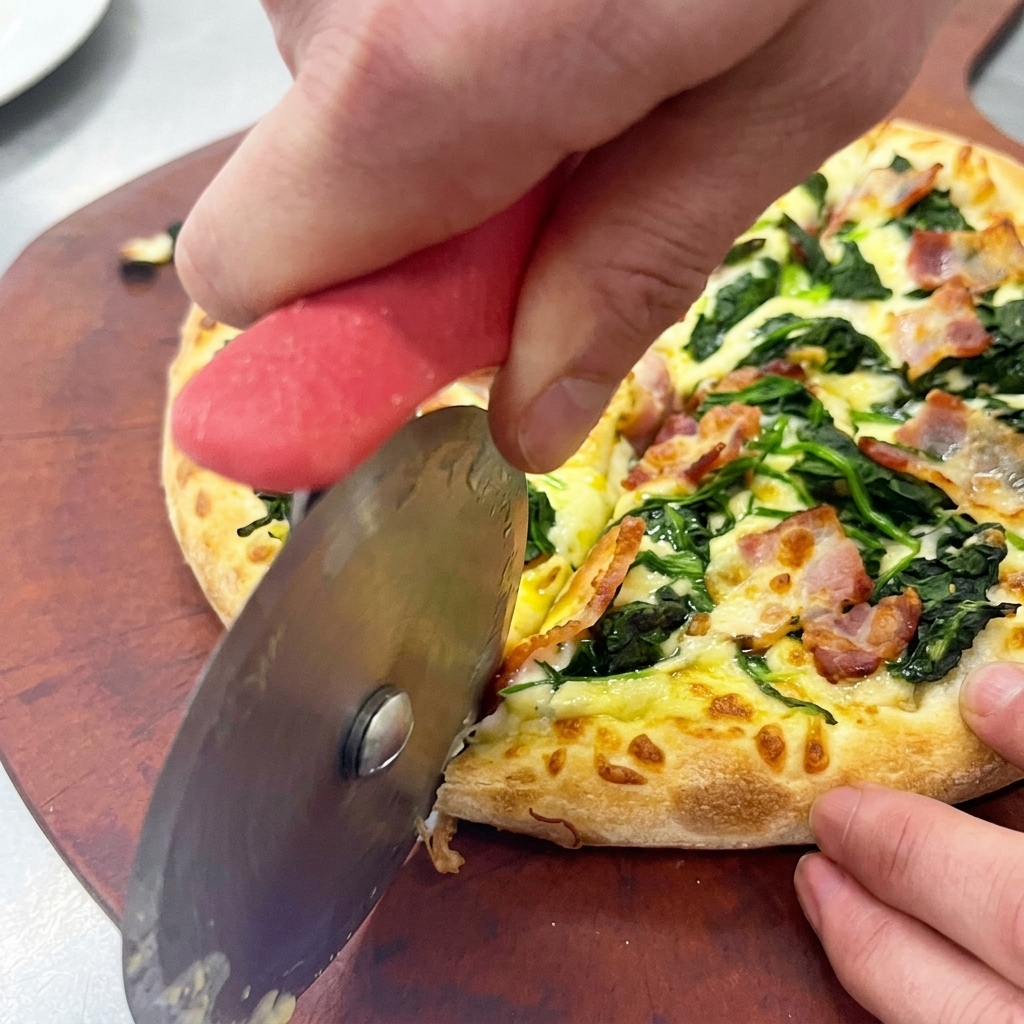 Hands using a rolling metal pizza cutter to slice a freshly baked spinach and bacon pizza on a round wooden peel.