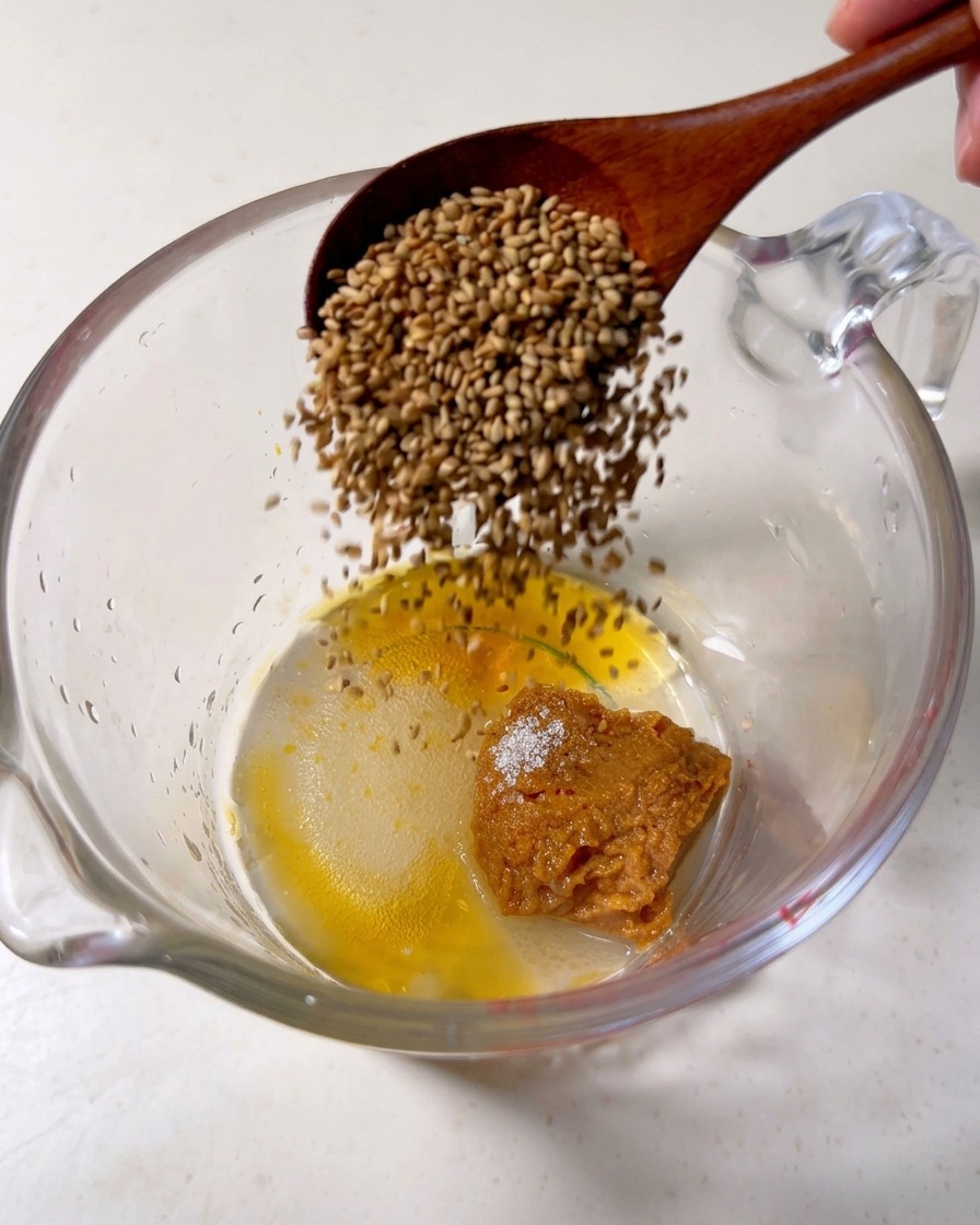 A wooden spoon pouring toasted sesame seeds into a glass measuring cup containing a dollop of miso paste, sugar, and sesame oil.