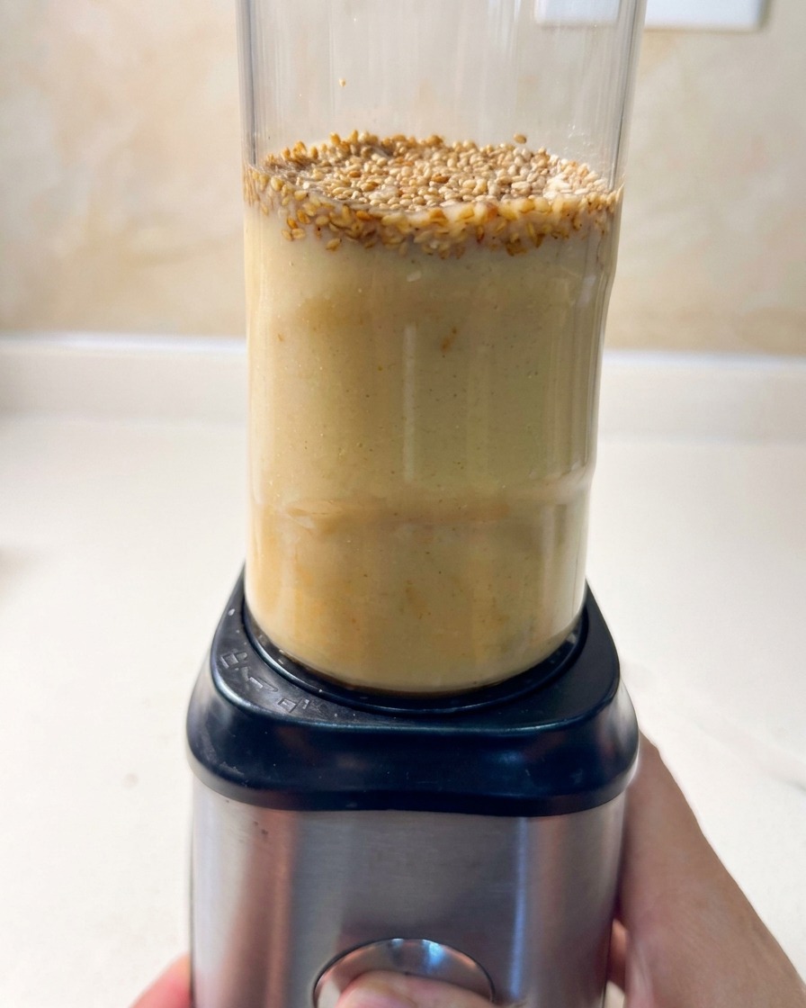 A hand turning the dial on a small personal blender filled with ramen broth ingredients and sesame seeds floating on top.