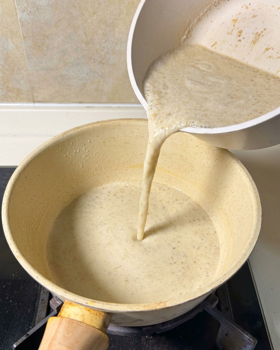 A smooth, blended beige soup mixture being poured from a white pitcher into a light yellow pot sitting on a stovetop.