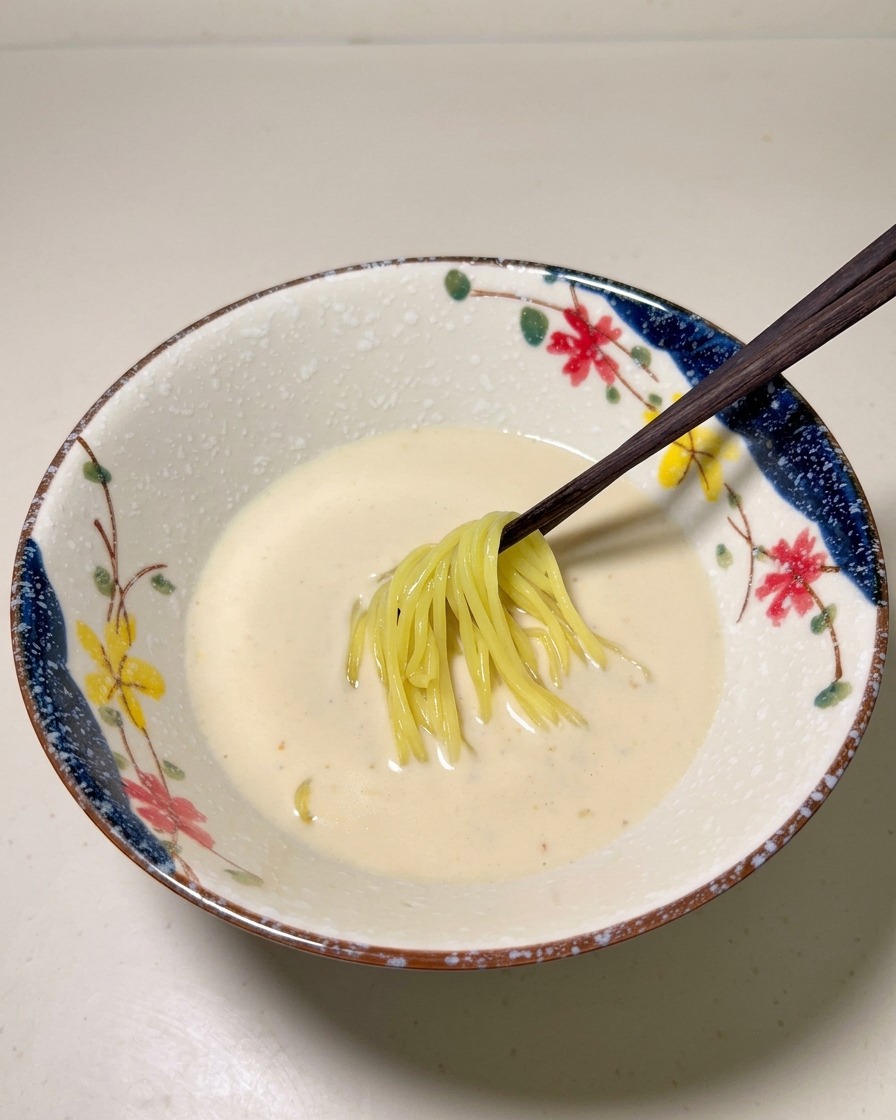 Cooked yellow ramen noodles being placed into a creamy, pale soup base in a decorative bowl.