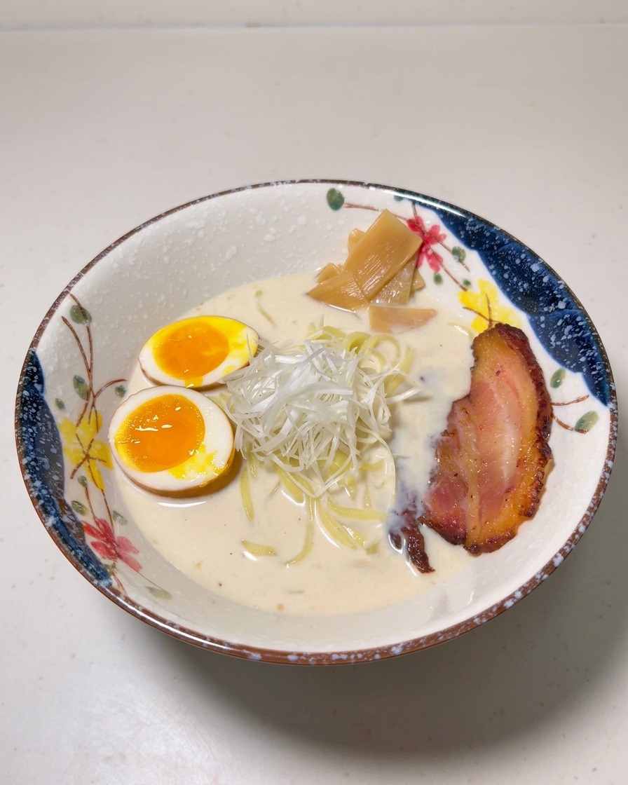 A ramen bowl topped with soft-boiled eggs, bamboo shoots, shredded white scallions, and a slice of browned chashu pork.