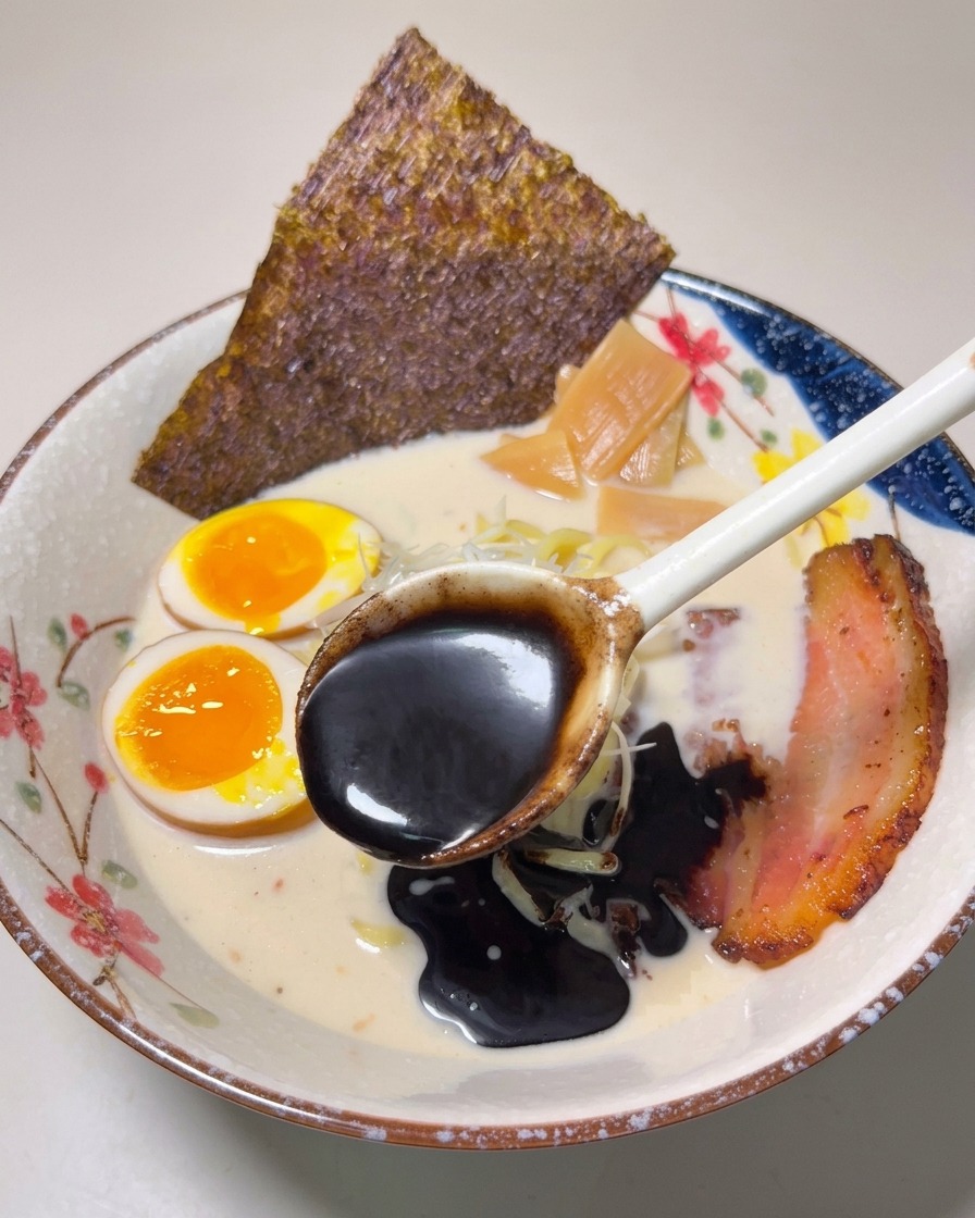 A spoonful of thick, dark black garlic oil being drizzled over a bowl of finished ramen.