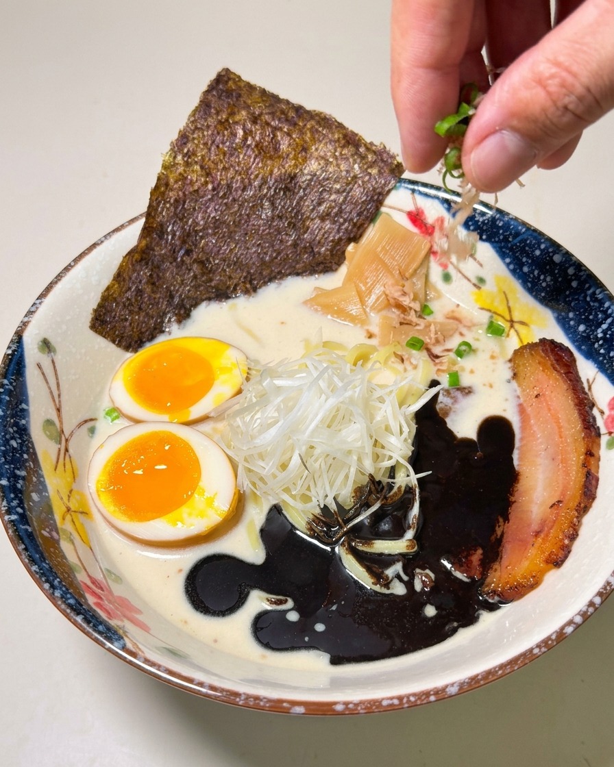 A hand sprinkling chopped scallions over a beautifully plated bowl of ramen featuring dark black garlic oil, chashu, and a soft-boiled egg.