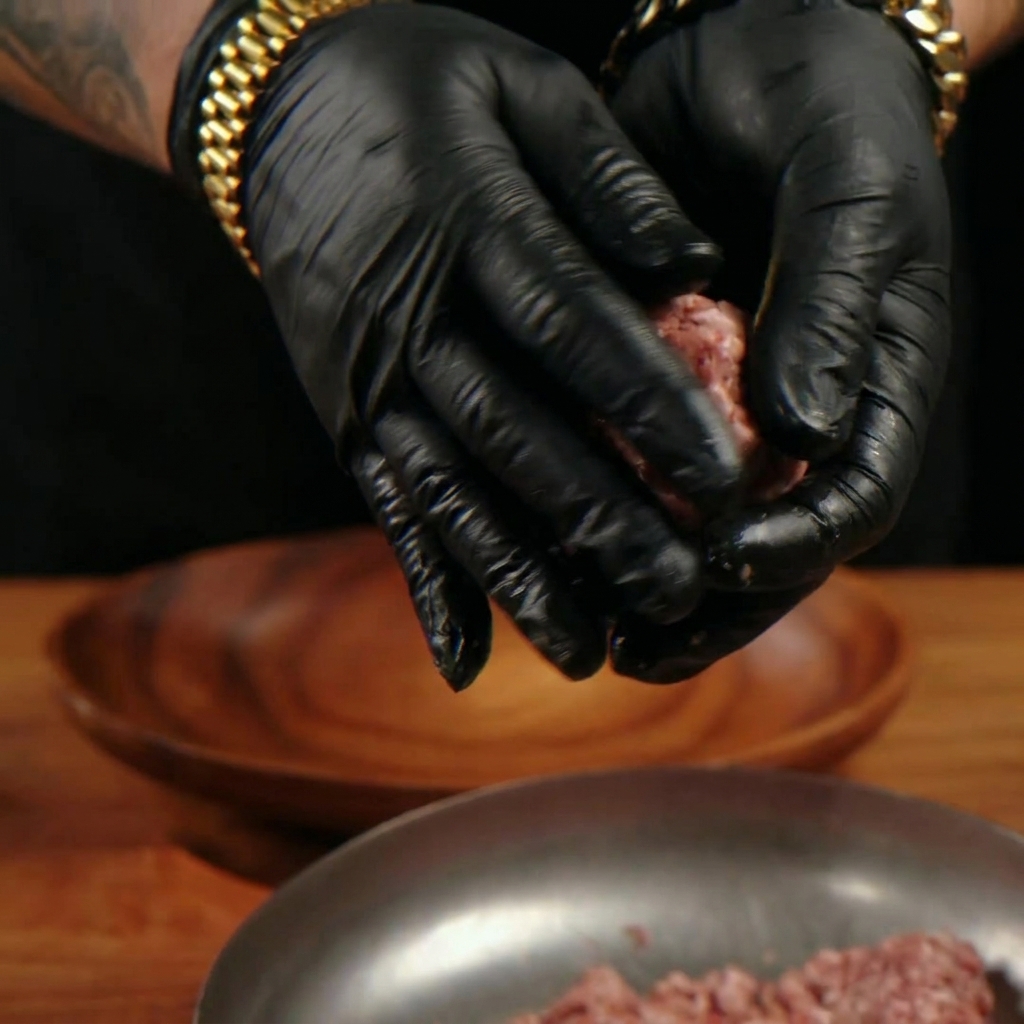 Gloved hands gently shaping raw ground beef into a round, loose patty over a wooden plate.