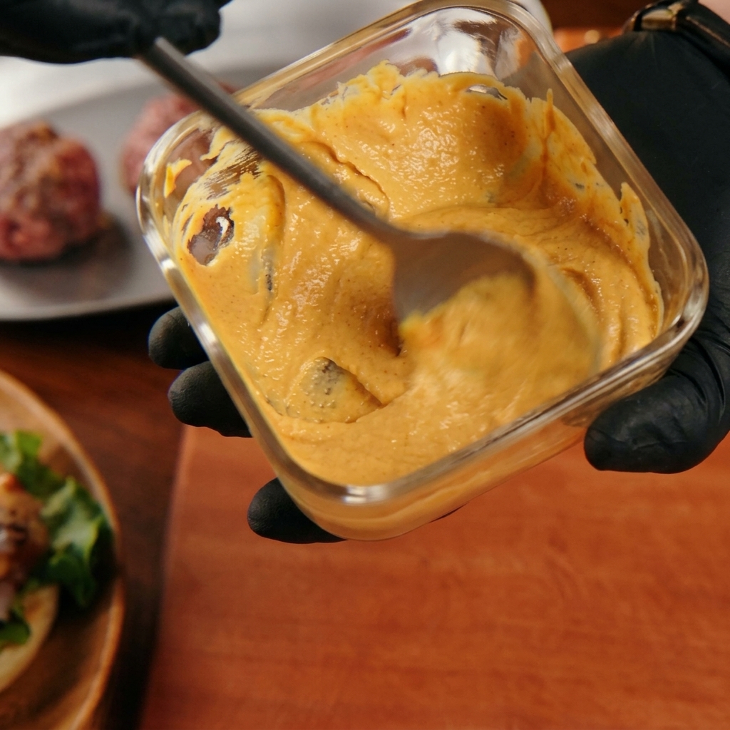A hand mixing a creamy orange burger sauce with a spoon in a square glass container.