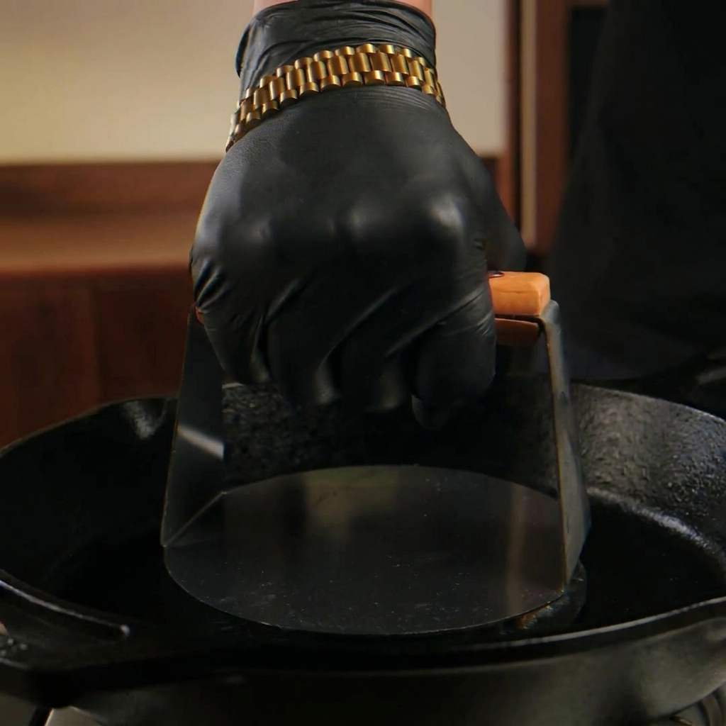 A hand wearing a black glove uses a heavy metal press to smash a raw beef ball flat onto a smoking hot cast-iron skillet.