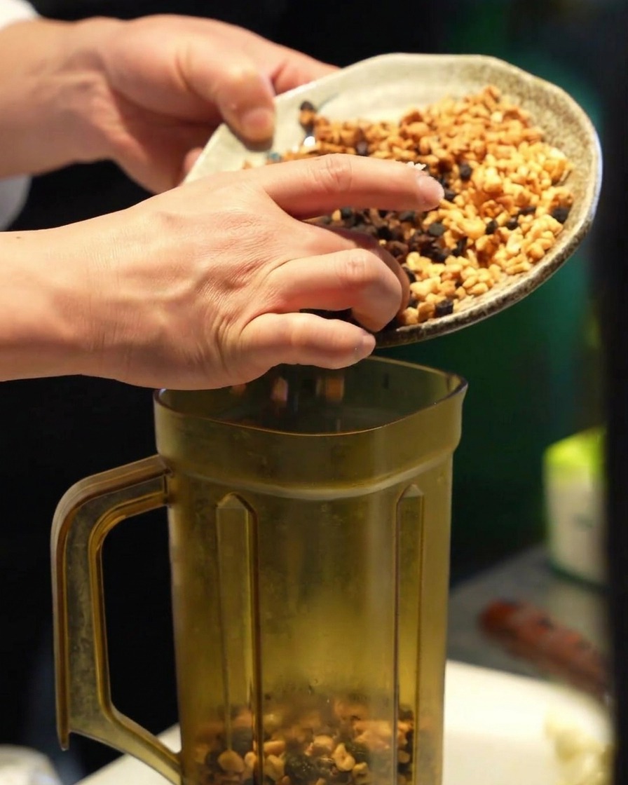 Hands scraping a colorful mixture of fried minced garlic into a tall, clear blender pitcher.