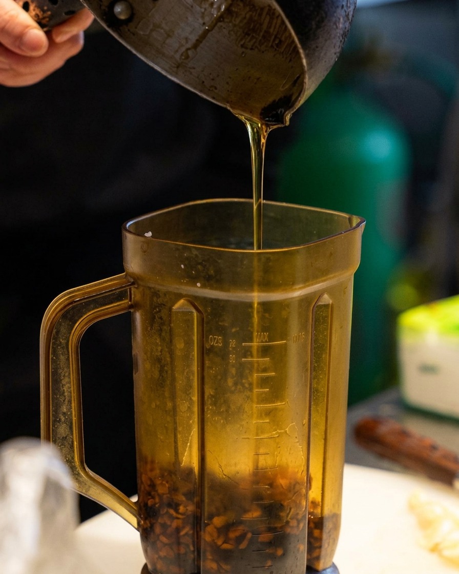 Pouring golden sesame oil from a small metal pot into a tall blender pitcher containing dark brown pieces of fried garlic.