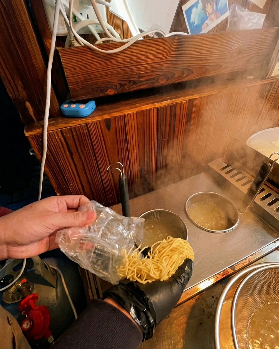Emptying fresh uncooked ramen noodles from a small plastic bag into a metal boiling basket submerged in a pot.