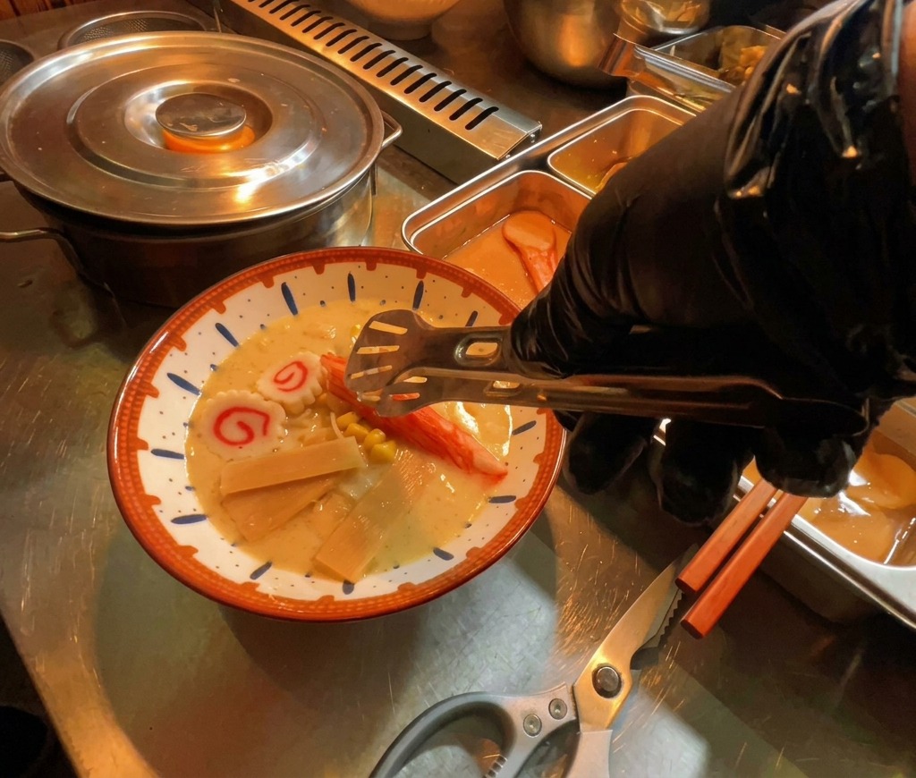 A gloved hand placing slices of narutomaki fish cake into a steaming bowl of ramen.