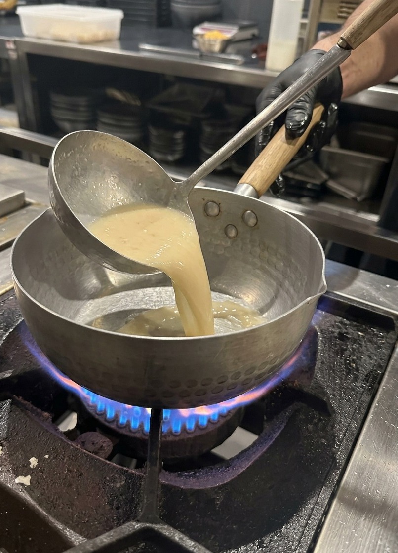 A metal ladle pouring light-colored pork bone broth into a heated metal pot on a gas stove.