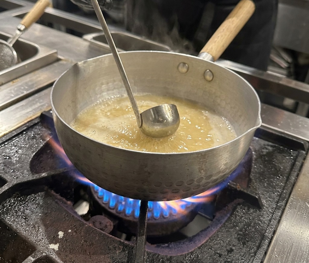 A gloved hand holding a small metal bowl, adding dark soy sauce into a bubbling pot of broth.