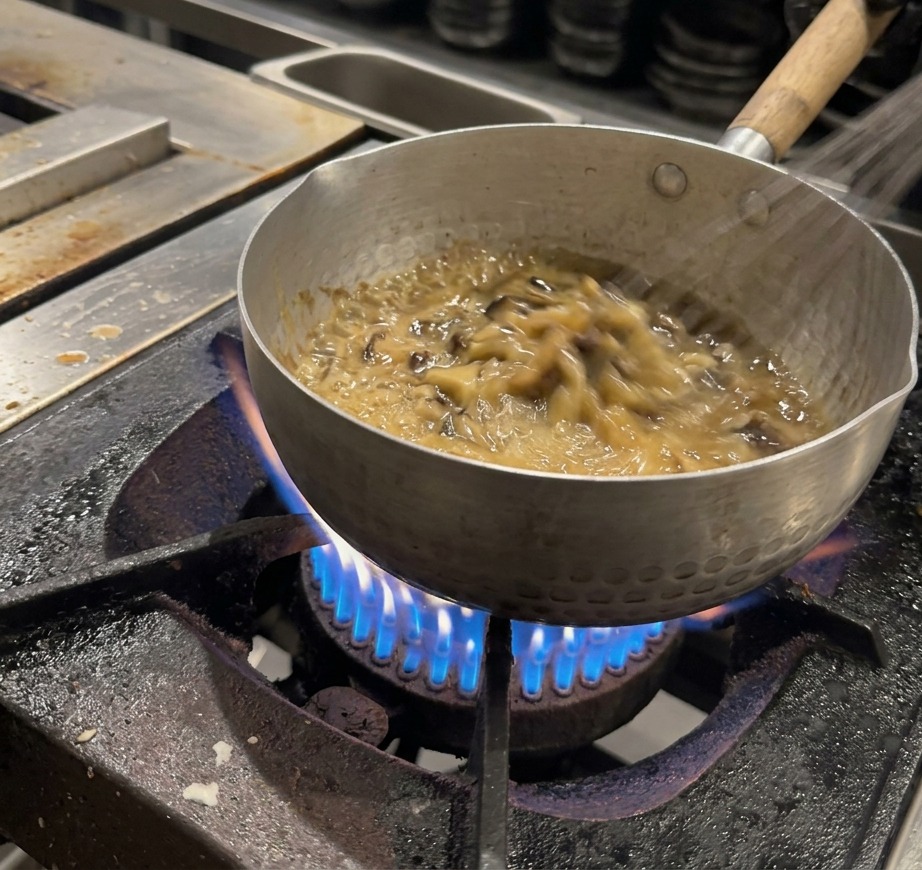 A wire whisk continuously stirring a thick, bubbling, dark brown sauce in a metal pot.