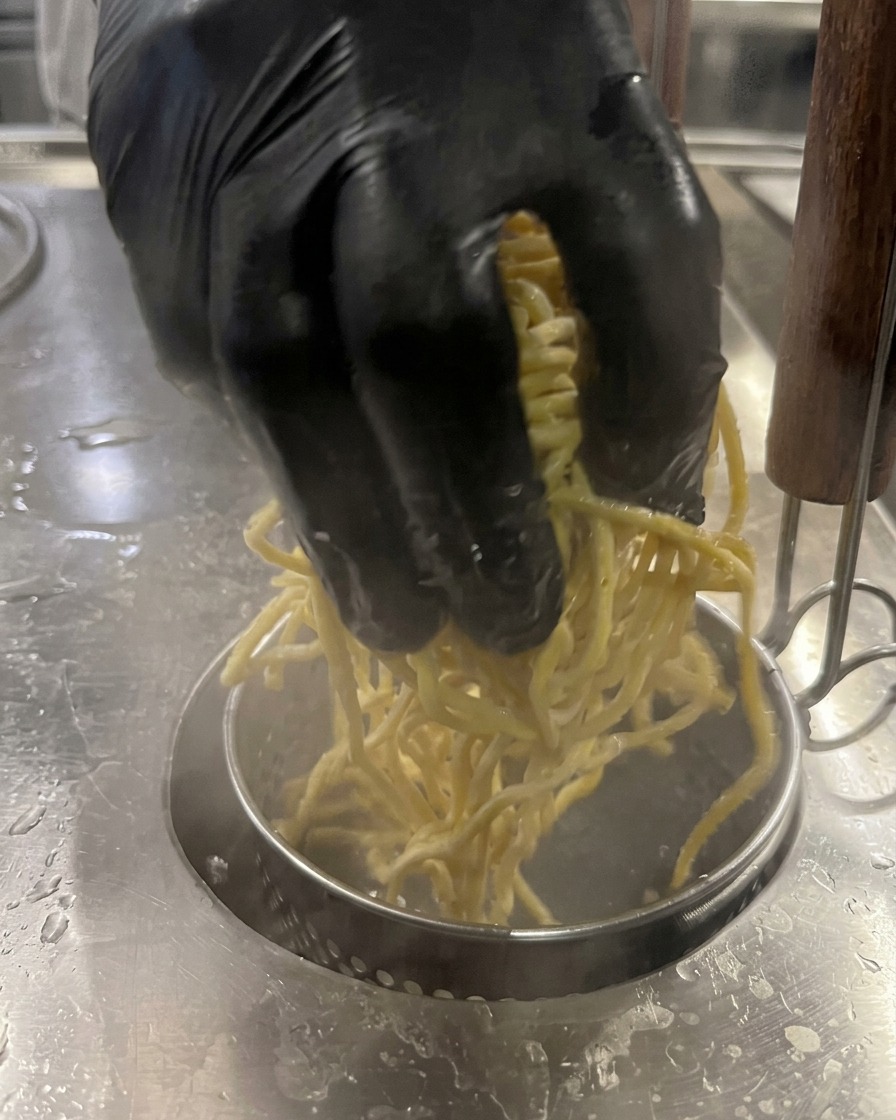 A gloved hand lowering a batch of yellow noodles into a pot of boiling water.