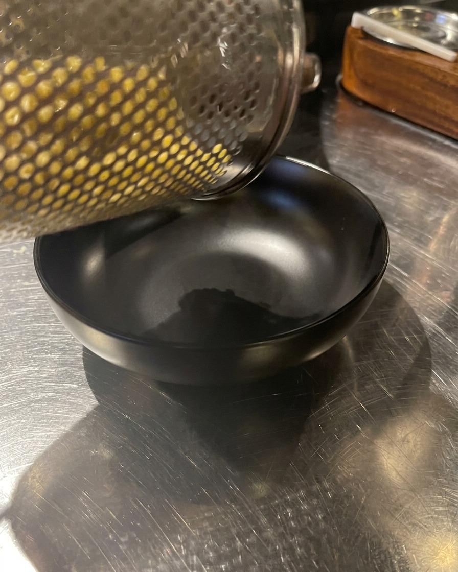 Freshly cooked yellow noodles being emptied from a metal strainer into a black bowl.