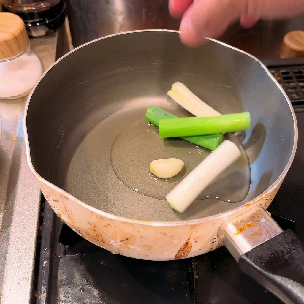 A hand dropping large pieces of chopped scallions and a clove of garlic into a cold pot filled with cooking oil.
