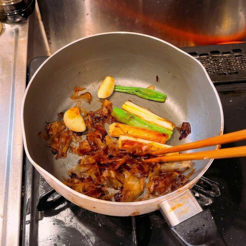 Chopsticks stirring heavily caramelized, dark brown scallions, garlic cloves, and bonito flakes in a pot over heat.