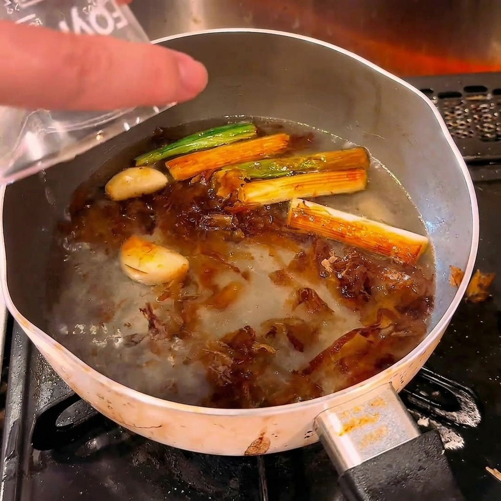Clear water being poured from a measuring cup into a hot pot containing browned scallions, garlic, and bonito flakes.