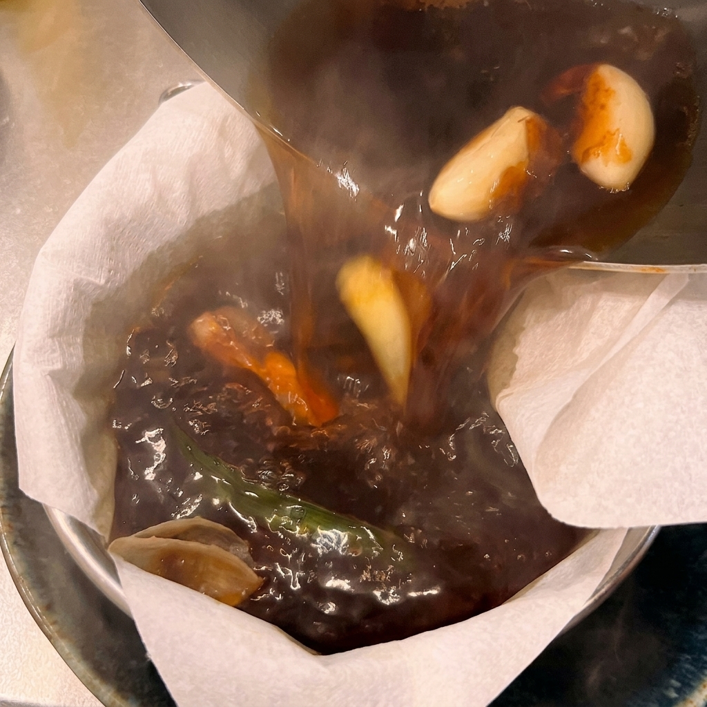 Dark, hot ramen broth with whole garlic cloves being poured from a pot through a white paper filter into a serving bowl.
