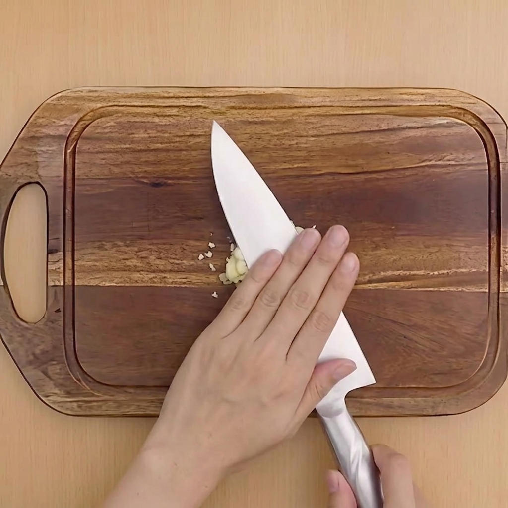 A hand pressing down on the flat side of a chefs knife to crush a garlic clove on a wooden cutting board.