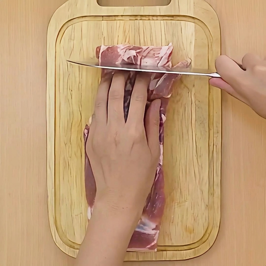 A hand holding a knife and making shallow horizontal cuts into a piece of raw pork belly on a cutting board.