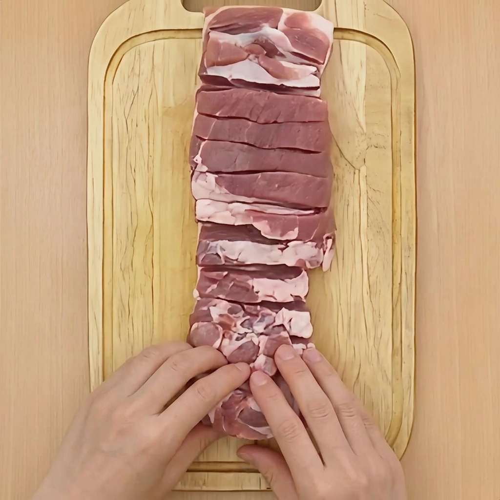 Two hands tightly rolling a block of raw pork belly into a cylindrical shape on a wooden board.