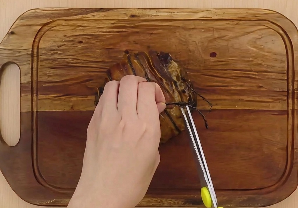 Hands using bright yellow-handled kitchen scissors to cut and remove dark butchers twine from a braised pork belly roll on a wooden cutting board.