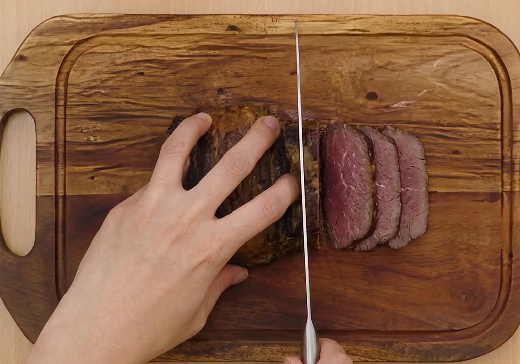 Hands firmly holding a braised pork belly roll while using a long, sharp carving knife to slice it into thin rounds on a wooden cutting board.