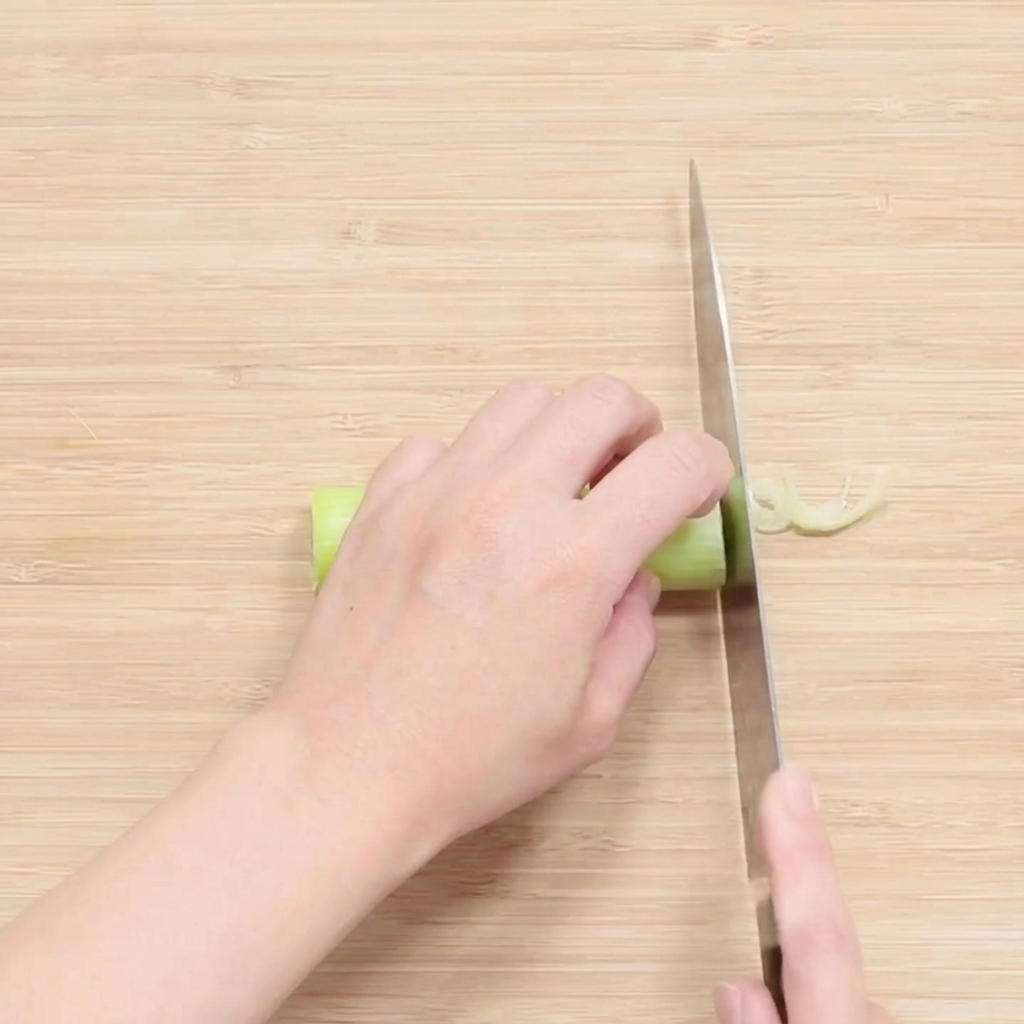 Hands using a large knife to thinly slice the white portion of a green onion on a wooden cutting board.