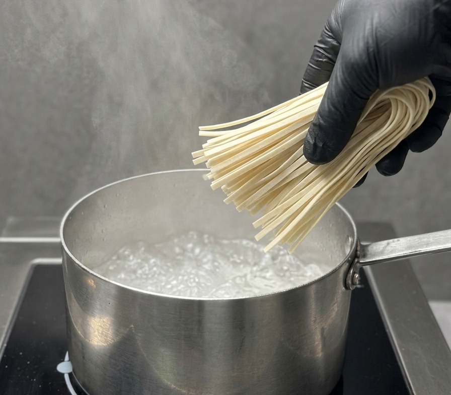A black-gloved hand carefully dropping a bundle of straight, dry ramen noodles into a stainless steel pot of rapidly boiling water.