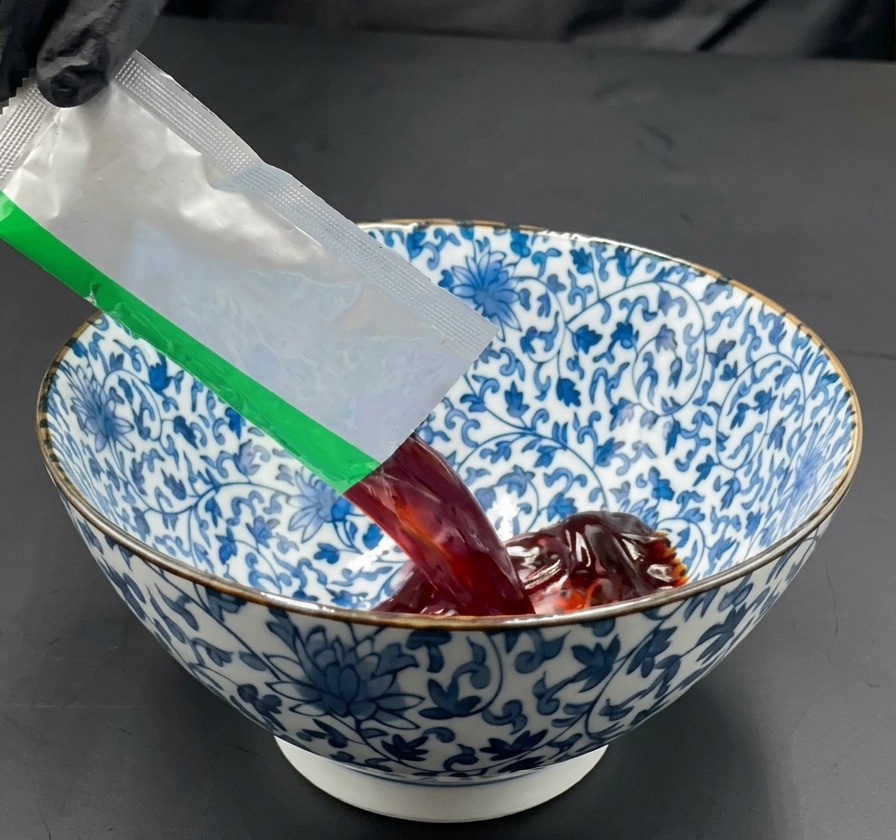 A hand pouring a packet of dark soy sauce soup base into a blue and white patterned ceramic bowl.