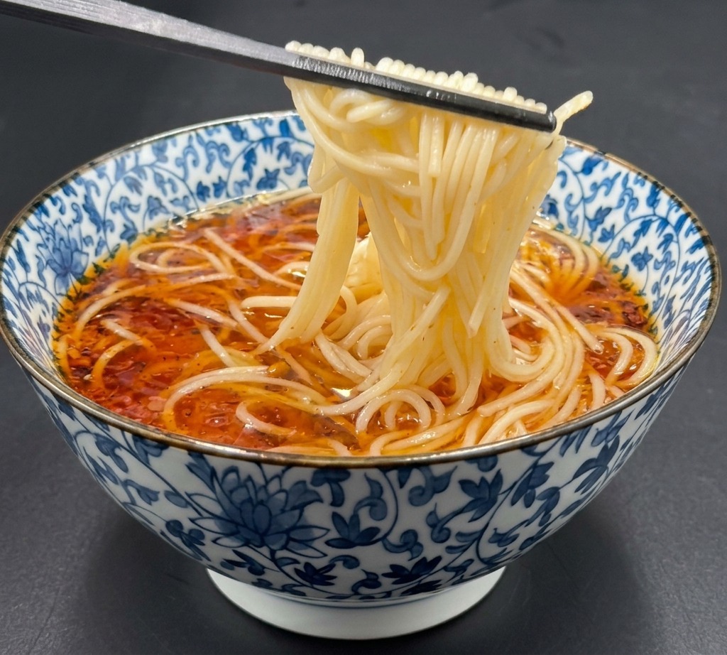 Chopsticks lifting cooked ramen noodles and placing them gently into a hot soy sauce broth in a patterned bowl.