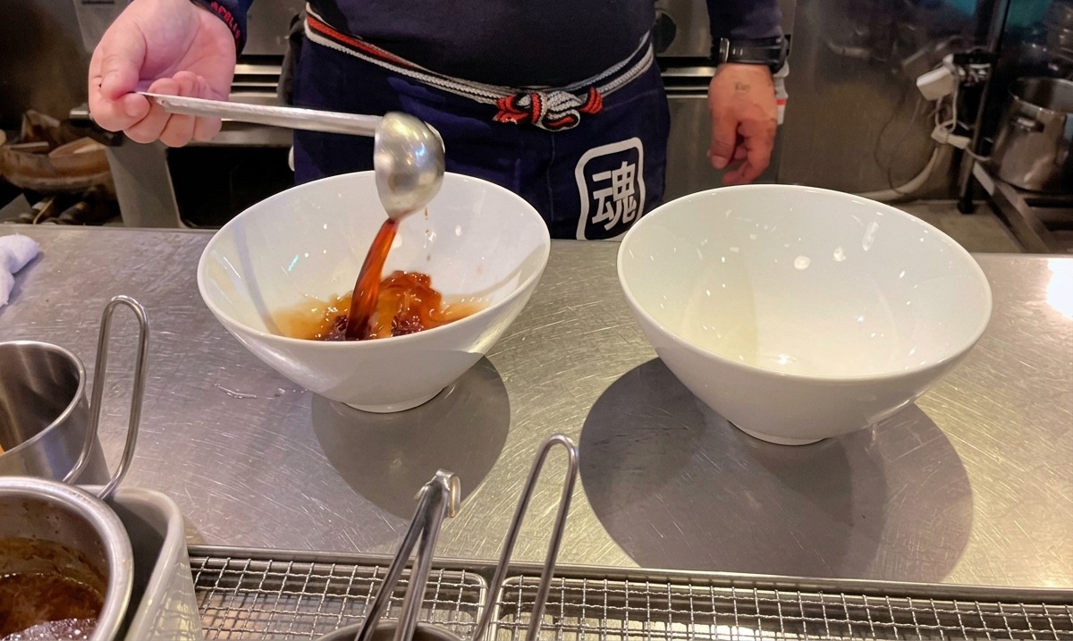 Chef pouring dark soy sauce tare into white ramen bowls using a ladle.