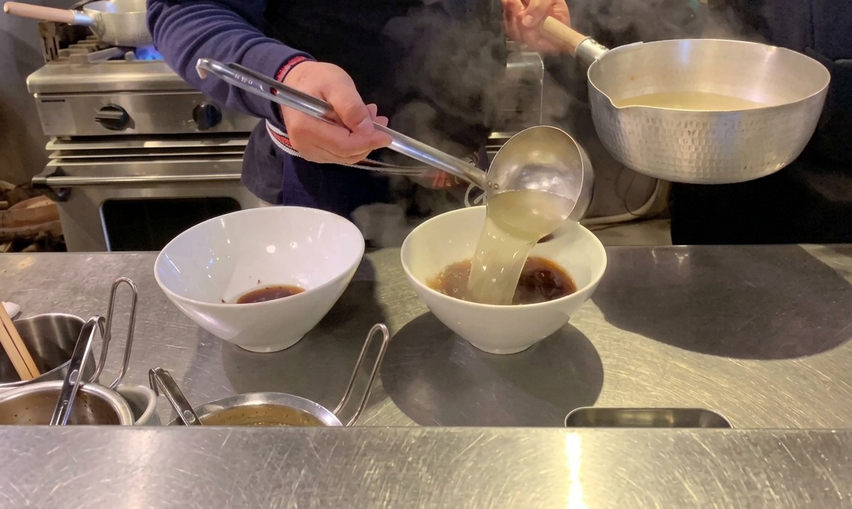 Pouring steaming clear hot broth into ramen bowls over dark soy sauce.