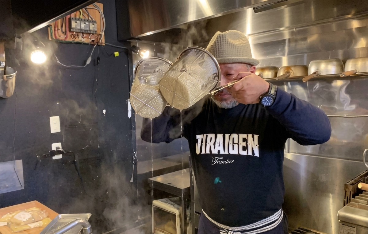 Chef vigorously shaking noodle strainers up high to drain water from cooked ramen.