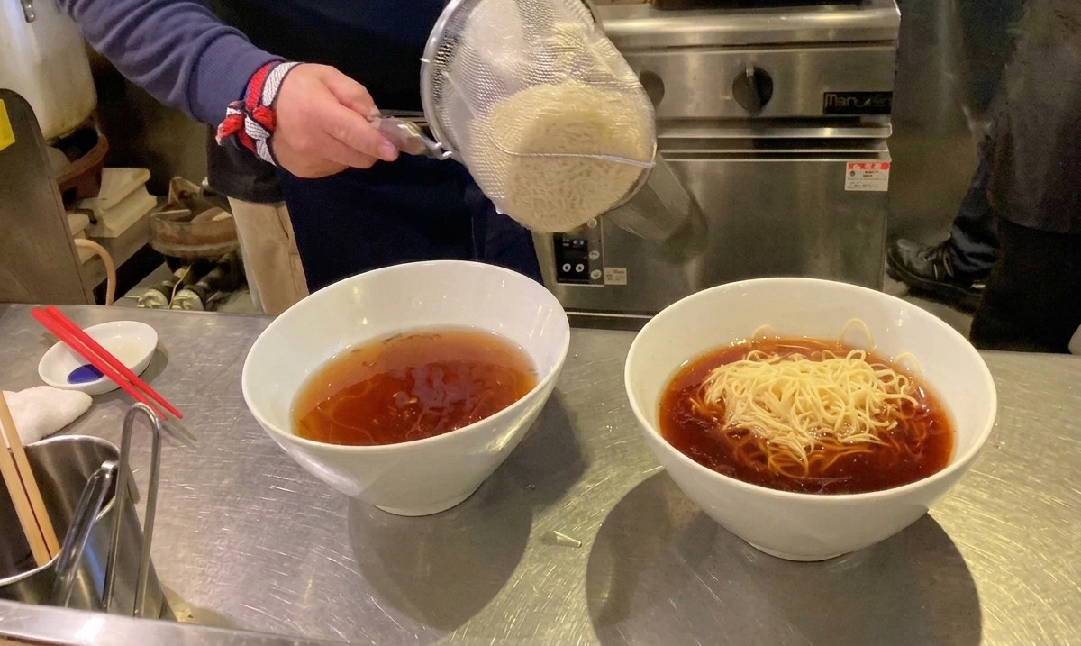 Freshly cooked ramen noodles being emptied from a strainer into a bowl of hot soup.