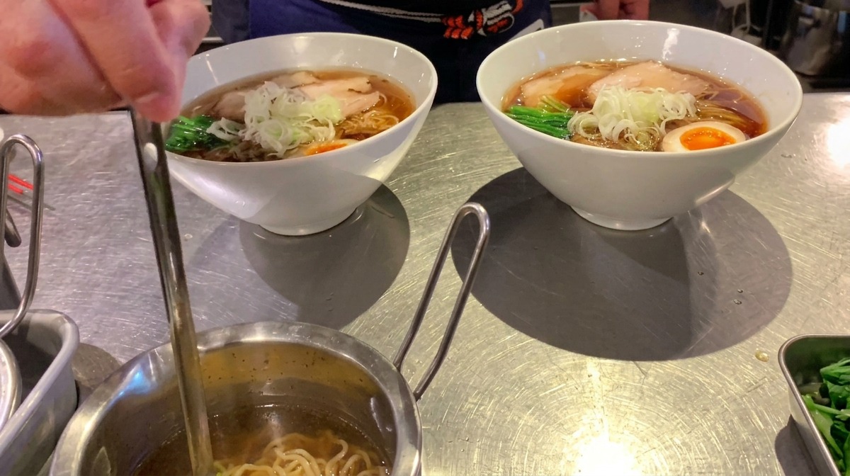 A small ladle drizzling clear aromatic oil over a finished bowl of ramen.