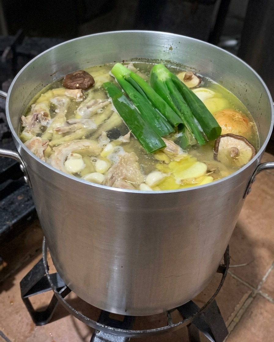 A large pot of boiling bone broth with whole green scallions, ginger slices, shiitake mushrooms, and apple halves added.