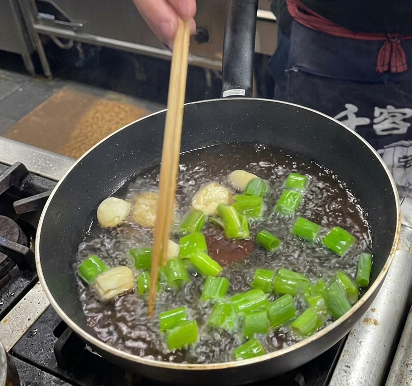 Frying large chunks of green scallions, garlic cloves, and ginger slices in hot rendered pork fat in a dark pan.