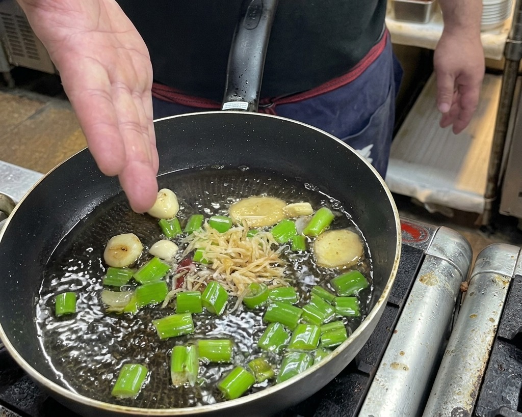A hand tossing dried shrimp into a frying pan filled with hot oil, ginger slices, garlic, and scallions.