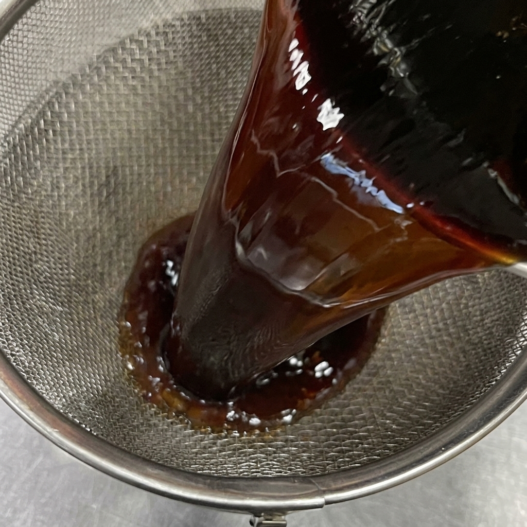 Dark, rich soy sauce mixture being poured through a fine-mesh metal sieve to remove braising ingredients.